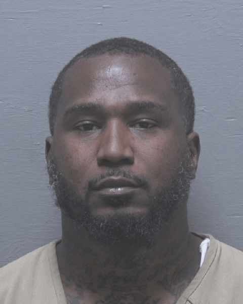 A man with short hair and a beard wearing a beige shirt stands against a plain gray background.