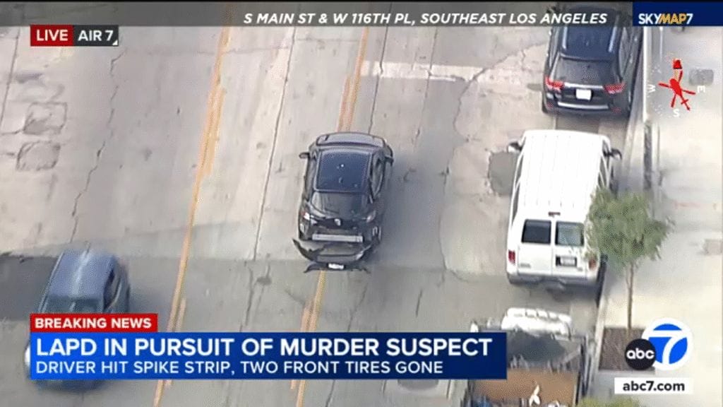A damaged black car is pursued by police vehicles on a street as news text reports an LAPD chase of a murder suspect with blown front tires in Southeast Los Angeles.