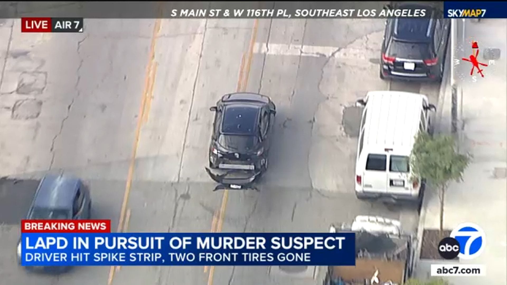 A damaged black car is pursued by police vehicles on a street as news text reports an LAPD chase of a murder suspect with blown front tires in Southeast Los Angeles.