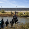 A group of people with backpacks wade through a shallow river toward a border wall topped with barbed wire under a clear sky.