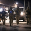 Five armed law enforcement officers in tactical gear walk together at night near a fenced area and parked vehicles.