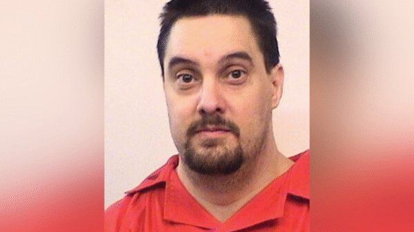 A man with short dark hair and a goatee, wearing a red shirt, looks at the camera against a plain, light-colored background.
