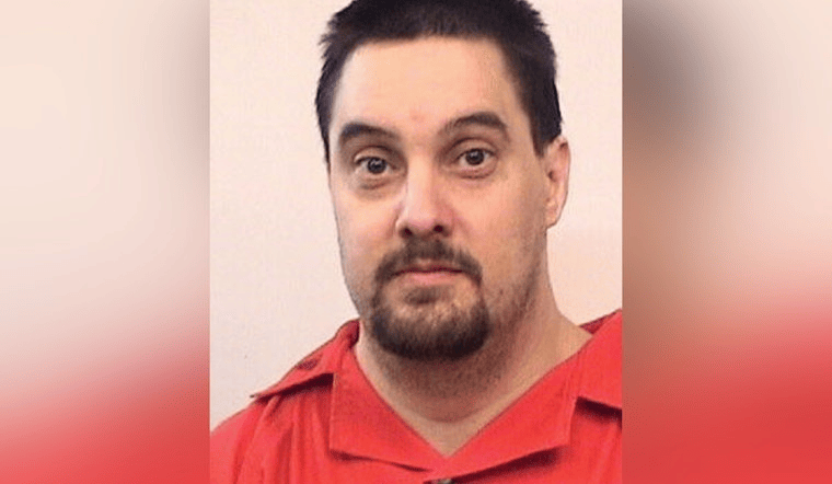 A man with short dark hair and a goatee, wearing a red shirt, looks at the camera against a plain, light-colored background.