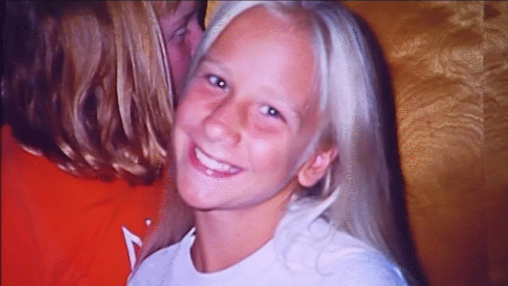 A young girl with long blonde hair smiles at the camera, wearing a white shirt. Another person with light brown hair is beside her, partially out of frame.