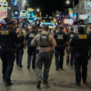 A group of police and ICE officers walk down a busy, neon-lit street at night, surrounded by crowds and vibrant signage.