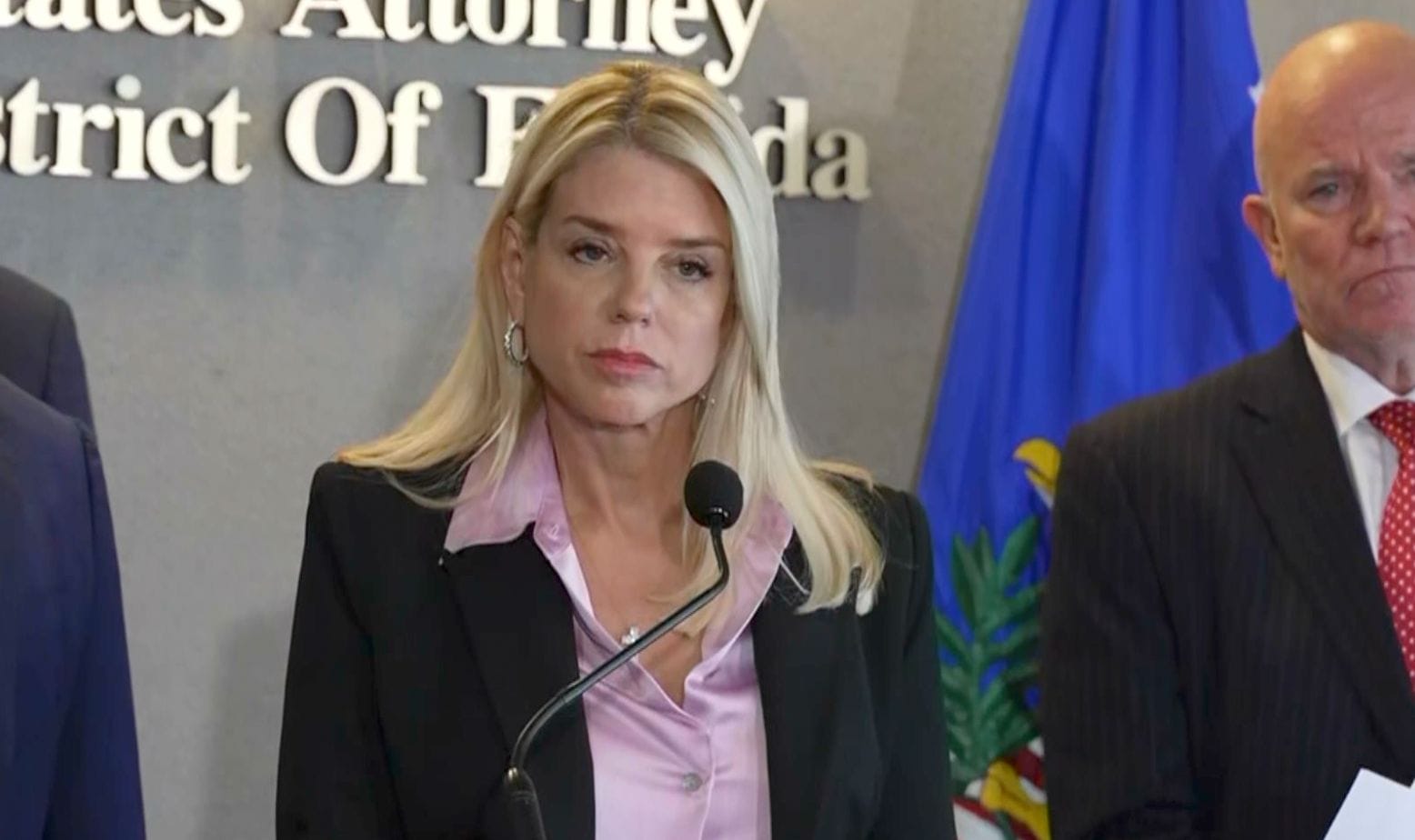 A woman in a black blazer and pink blouse speaks at a podium with a microphone, standing in front of a wall with text and beside a man in a suit.