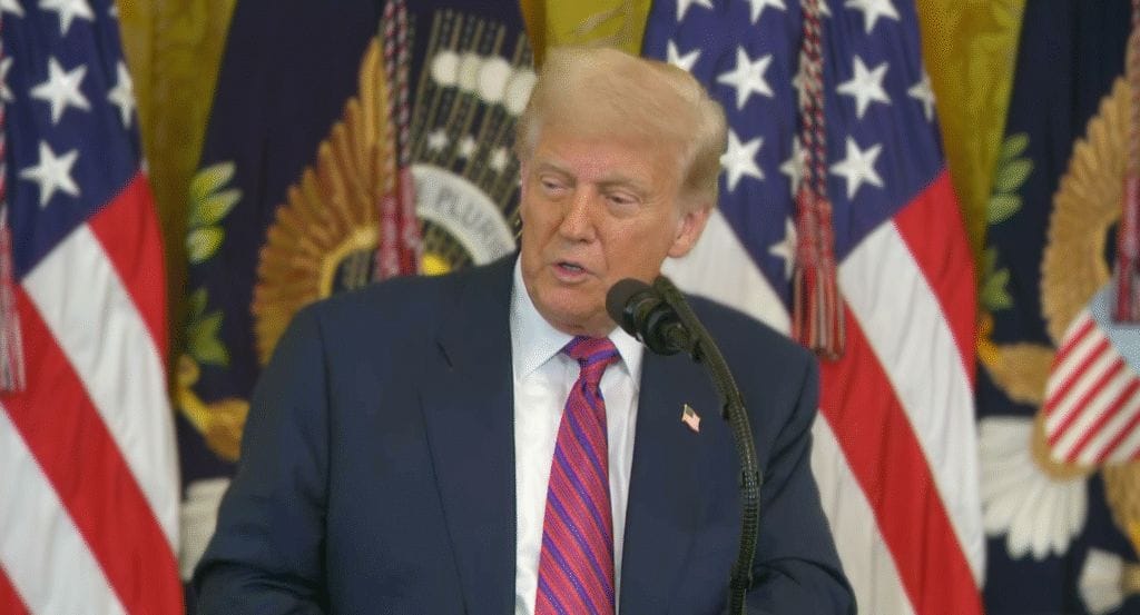 A man in a suit and red striped tie speaks at a podium, with U.S. flags and decorative banners in the background.