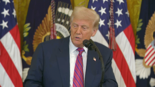 A man in a suit and red striped tie speaks at a podium, with U.S. flags and decorative banners in the background.