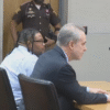 A man and his attorney sit at a courtroom table, while a police officer and two deputies stand in the background.