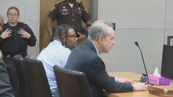 A man and his attorney sit at a courtroom table, while a police officer and two deputies stand in the background.