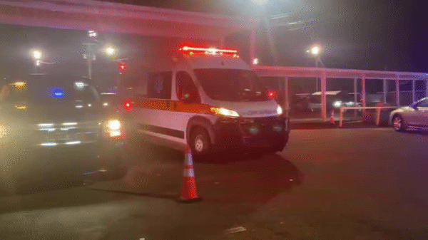 An ambulance with flashing red lights is parked at night next to other vehicles, with an orange traffic cone in front of it and bright lights illuminating the scene.