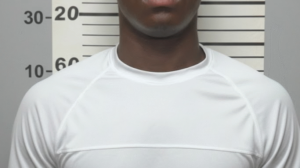 A young man in a white shirt stands in front of a height measurement chart, posing for a mugshot-style photo.