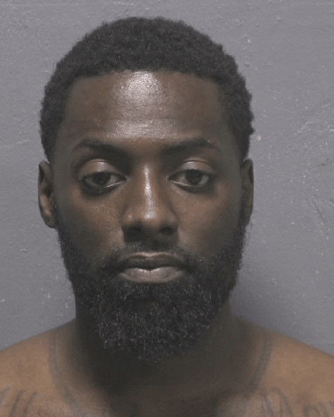 A man with short curly hair, a full beard, and no shirt poses against a plain gray background.