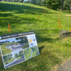 A sign for the Broome County Crime Victims Memorial stands on grass near a walking path, with orange and blue stakes marking spots on the lawn in the background.