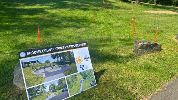 A sign for the Broome County Crime Victims Memorial stands on grass near a walking path, with orange and blue stakes marking spots on the lawn in the background.