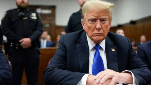 A man in a suit and blue tie sits at a courtroom table, looking forward, with police officers and people in the background.