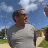 A man in handcuffs stands outside in a neighborhood while a police officer talks to him under a blue sky with scattered clouds.