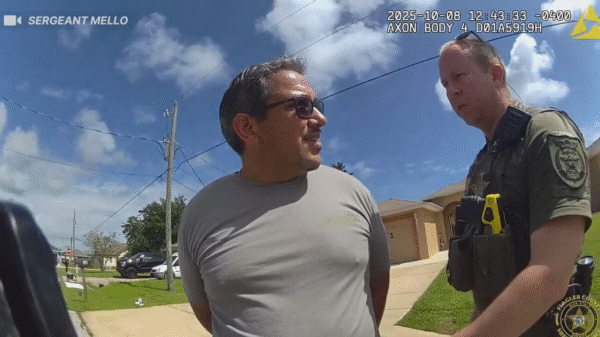 A man in handcuffs stands outside in a neighborhood while a police officer talks to him under a blue sky with scattered clouds.