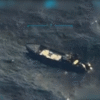 Aerial view of a dark-colored ship sailing in the ocean, seen through a targeting or surveillance camera interface.