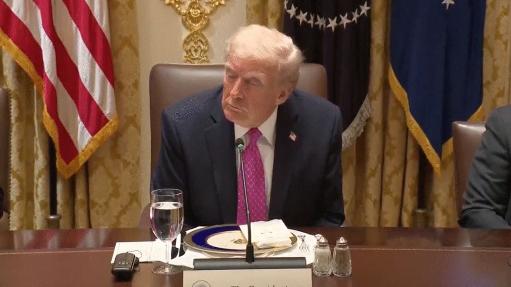 A man in a suit and pink tie sits at a formal dining table with a glass of water, plates, and documents in front of him, with U.S. flags and gold curtains in the background.