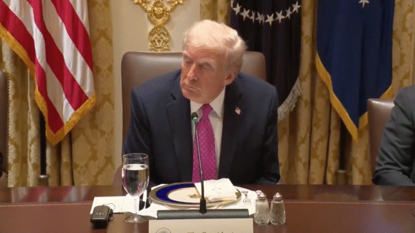 A man in a suit and pink tie sits at a formal dining table with a glass of water, plates, and documents in front of him, with U.S. flags and gold curtains in the background.