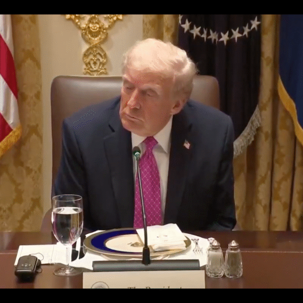 A man in a suit and pink tie sits at a formal dining table with a glass of water, plates, and documents in front of him, with U.S. flags and gold curtains in the background.