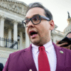 A person in a maroon suit, red tie, and glasses speaks to reporters outside a government building with columns and a dome in the background.