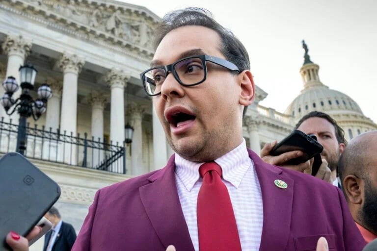 A person in a maroon suit, red tie, and glasses speaks to reporters outside a government building with columns and a dome in the background.