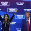 A woman raises both arms and smiles while standing next to a man in a suit and red tie, with a blue "Trump Vance" campaign backdrop behind them.