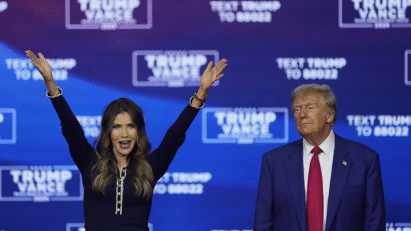 A woman raises both arms and smiles while standing next to a man in a suit and red tie, with a blue "Trump Vance" campaign backdrop behind them.