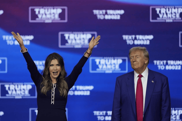 A woman raises both arms and smiles while standing next to a man in a suit and red tie, with a blue "Trump Vance" campaign backdrop behind them.