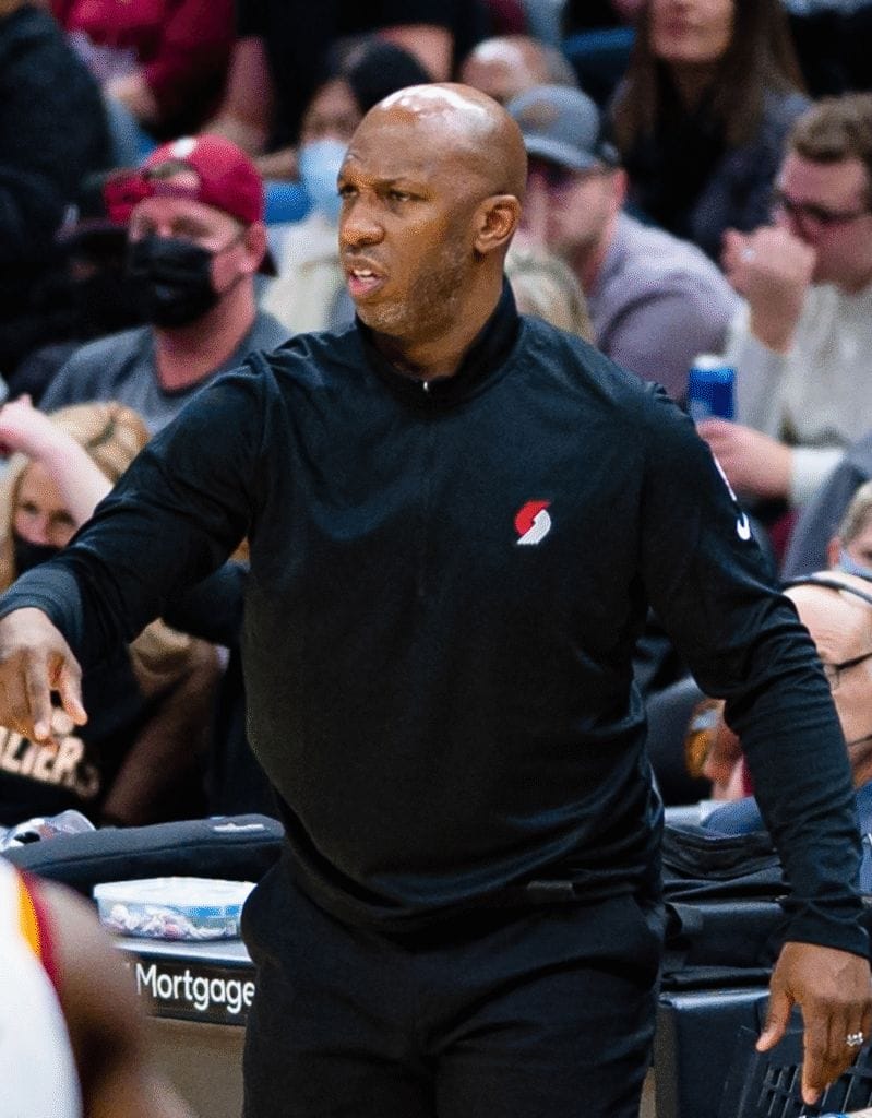 A basketball coach in a black zip-up jacket with a red and white logo stands courtside, gesturing with a focused expression during a game.