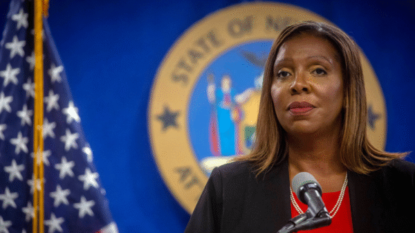 A woman stands at a podium with a microphone, wearing a red blouse and black jacket, in front of a U.S. flag and a blue and gold state seal.