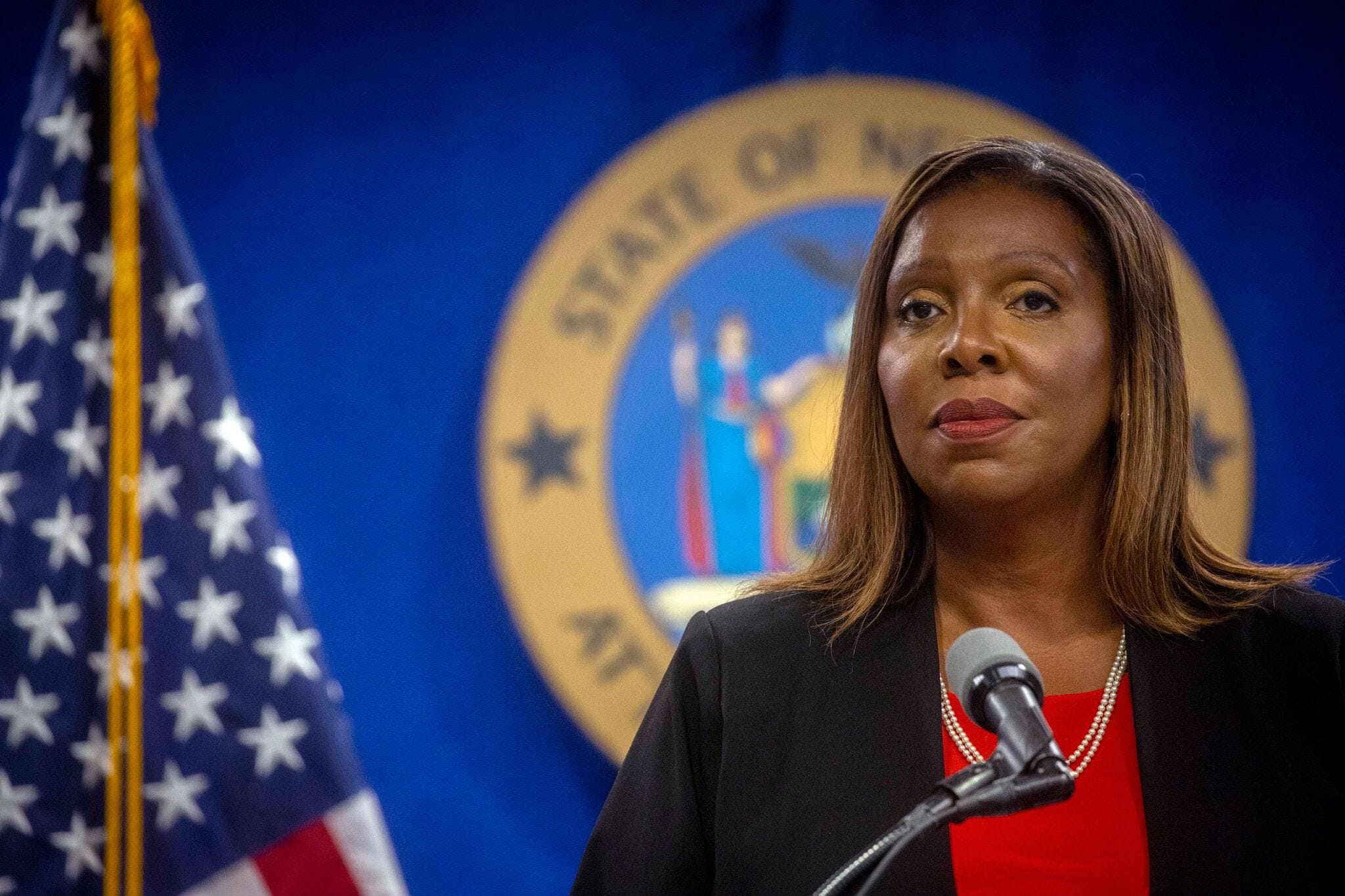 A woman stands at a podium with a microphone, wearing a red blouse and black jacket, in front of a U.S. flag and a blue and gold state seal.