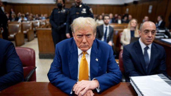 A man in a blue suit and yellow tie sits at a courtroom table, surrounded by people, with police officers and others in the background.