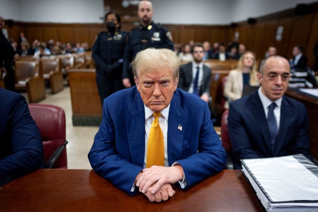 A man in a blue suit and yellow tie sits at a courtroom table, surrounded by people, with police officers and others in the background.
