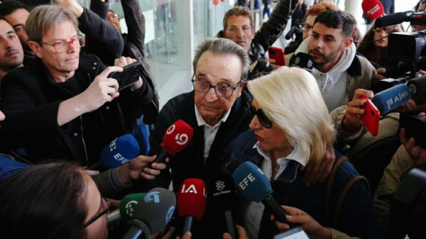 A man and woman surrounded by reporters and microphones, answering questions in a crowded indoor setting with cameras pointed at them.