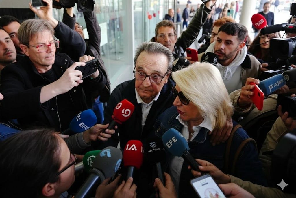 A man and woman surrounded by reporters and microphones, answering questions in a crowded indoor setting with cameras pointed at them.