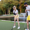 A woman and a man in sporty clothes play street hockey on a grassy area near a pond and fountain, surrounded by trees.
