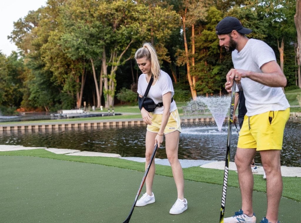 A woman and a man in sporty clothes play street hockey on a grassy area near a pond and fountain, surrounded by trees.