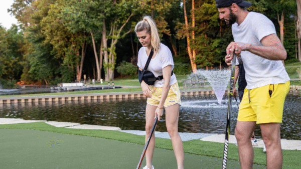 A woman and a man in sporty clothes play street hockey on a grassy area near a pond and fountain, surrounded by trees.