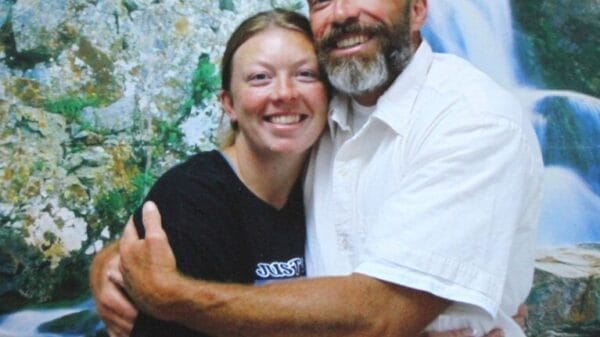 A smiling man and woman hug each other in front of a scenic waterfall and rocky landscape backdrop.