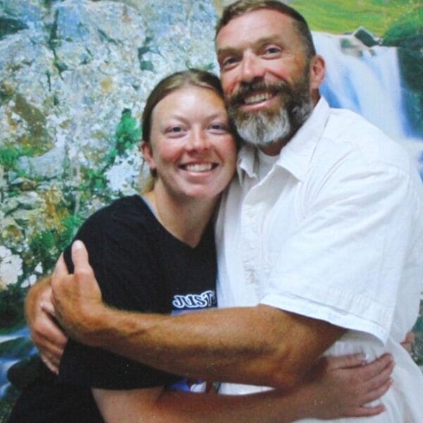 A smiling man and woman hug each other in front of a scenic waterfall and rocky landscape backdrop.