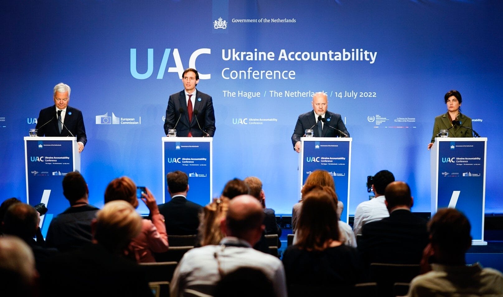 Four speakers stand at podiums on stage at the Ukraine Accountability Conference in The Hague, addressing an audience, with a blue backdrop displaying event details and logos.