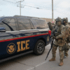 Armed officers in camouflage gear stand near an ICE vehicle, with a large group of similarly dressed personnel gathered in the background on a street.