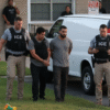 Four ICE officers detain a handcuffed man outside a house near a white van, with another man and a child visible in the background.