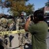 A man takes a photo of armed officers in camouflage behind yellow "Police Line Do Not Cross" tape on a city street during daytime.