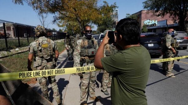A man takes a photo of armed officers in camouflage behind yellow "Police Line Do Not Cross" tape on a city street during daytime.