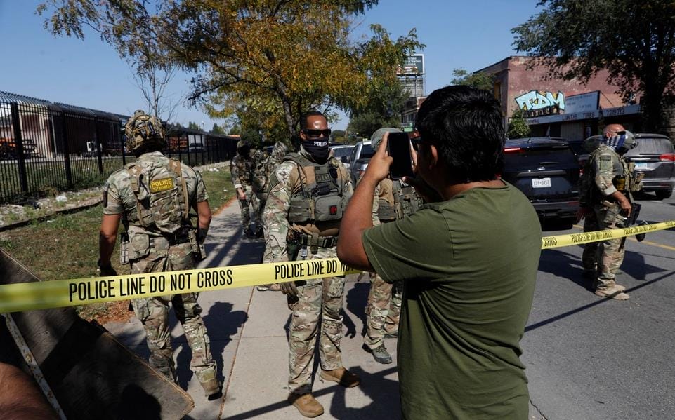 A man takes a photo of armed officers in camouflage behind yellow "Police Line Do Not Cross" tape on a city street during daytime.