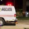 A Cleveland Police van is parked on a city street at night, with orange traffic cones nearby and a blurred blue car passing in the foreground.